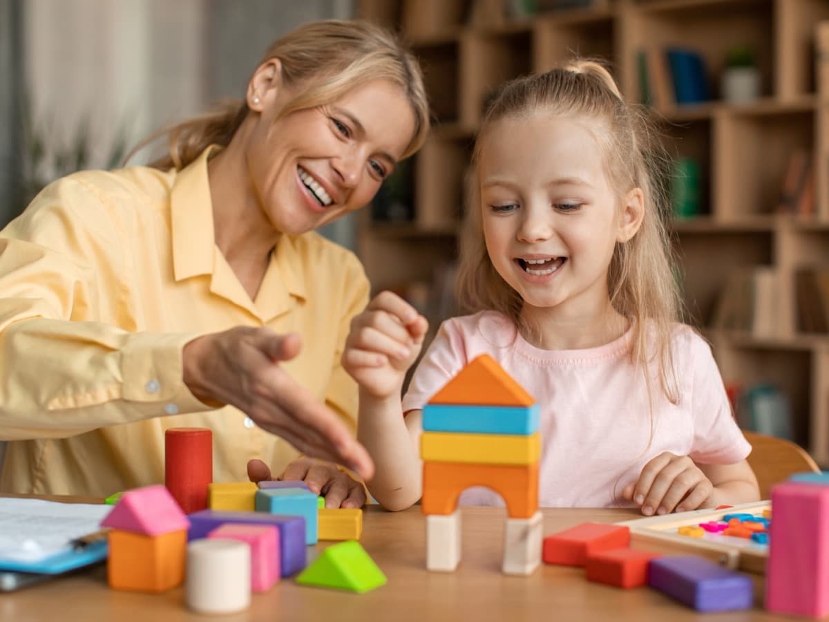 A woman and a young girl laugh together while stacking colorful wooden blocks at a low table.