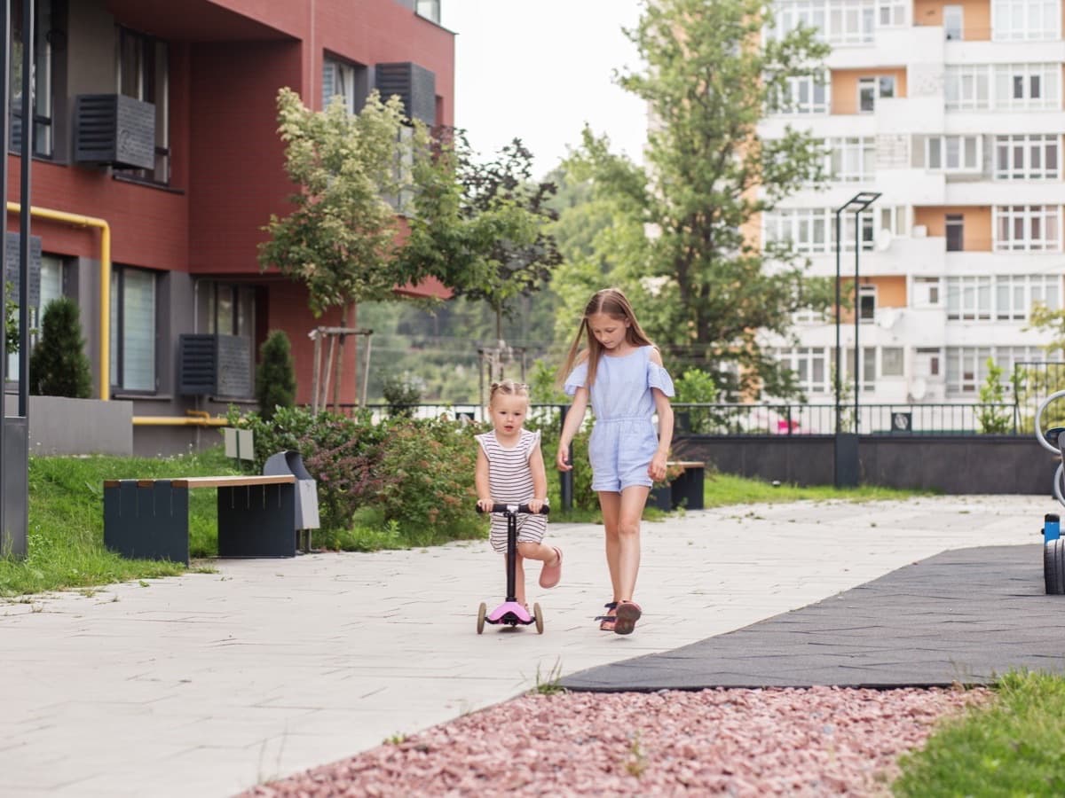 Two children make their way along a neighborhood sidewalk, the younger one on a scooter.